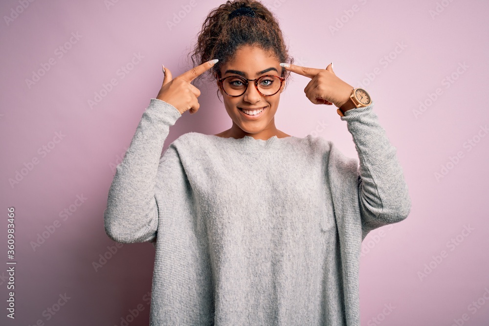 Young beautiful african american girl wearing sweater and glasses over pink background smiling pointing to head with both hands finger, great idea or thought, good memory Young beautiful african american girl wearing sweater and glasses over pink background smiling pointing to head with both hands finger, great idea or thought, good memory