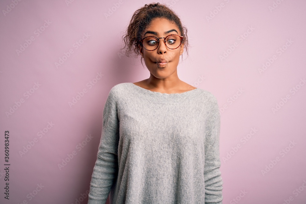Young beautiful african american girl wearing sweater and glasses over pink background making fish face with lips, crazy and comical gesture. Funny expression. Young beautiful african american girl wearing sweater and glasses over pink background making fish face with lips, crazy and comical gesture. Funny expression.