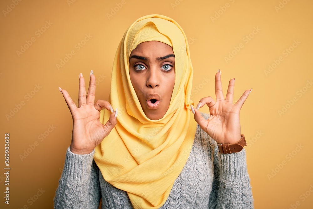 Young beautiful african american girl wearing muslim hijab over isolated yellow background looking surprised and shocked doing ok approval symbol with fingers. Crazy expression Young beautiful african american girl wearing muslim hijab over isolated yellow background looking surprised and shocked doing ok approval symbol with fingers. Crazy expression