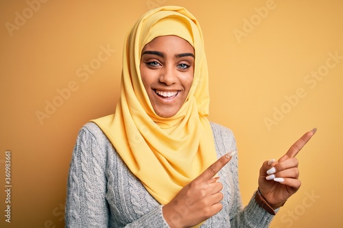 Young beautiful african american girl wearing muslim hijab over isolated yellow background smiling and looking at the camera pointing with two hands and fingers to the side. © Krakenimages.com