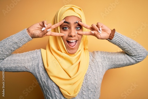 Young beautiful african american girl wearing muslim hijab over isolated yellow background Doing peace symbol with fingers over face, smiling cheerful showing victory