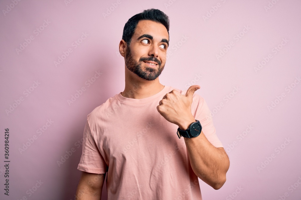 Young handsome man with beard wearing casual t-shirt standing over pink background smiling with happy face looking and pointing to the side with thumb up. Young handsome man with beard wearing casual t-shirt standing over pink background smiling with happy face looking and pointing to the side with thumb up.