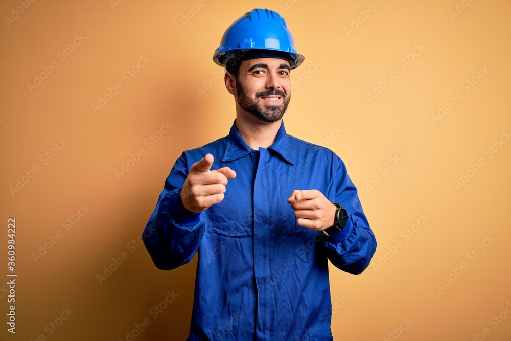 Mechanic man with beard wearing blue uniform and safety helmet over yellow background pointing fingers to camera with happy and funny face. Good energy and vibes.