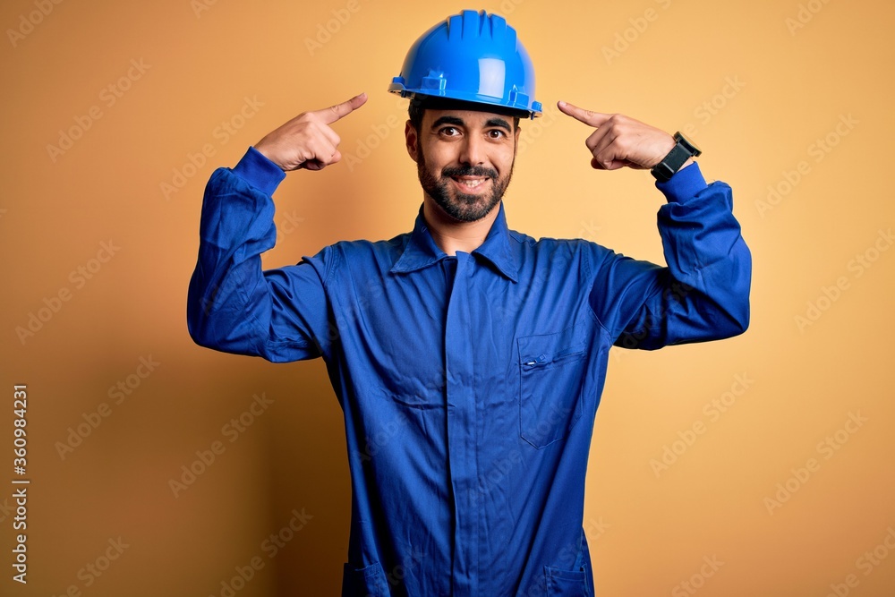 Mechanic man with beard wearing blue uniform and safety helmet over yellow background smiling pointing to head with both hands finger, great idea or thought, good memory