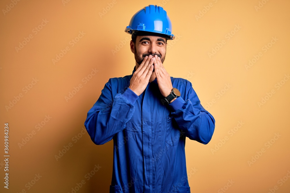 Mechanic man with beard wearing blue uniform and safety helmet over yellow background laughing and embarrassed giggle covering mouth with hands, gossip and scandal concept Mechanic man with beard wearing blue uniform and safety helmet over yellow background laughing and embarrassed giggle covering mouth with hands, gossip and scandal concept