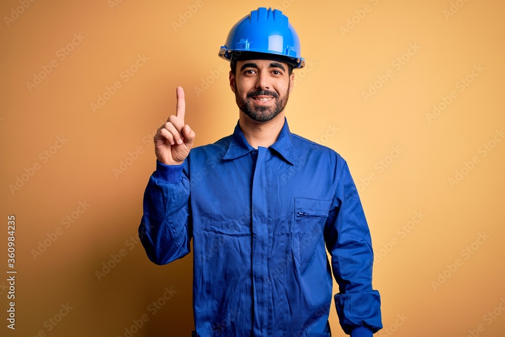 Mechanic man with beard wearing blue uniform and safety helmet over yellow background showing and pointing up with finger number one while smiling confident and happy.