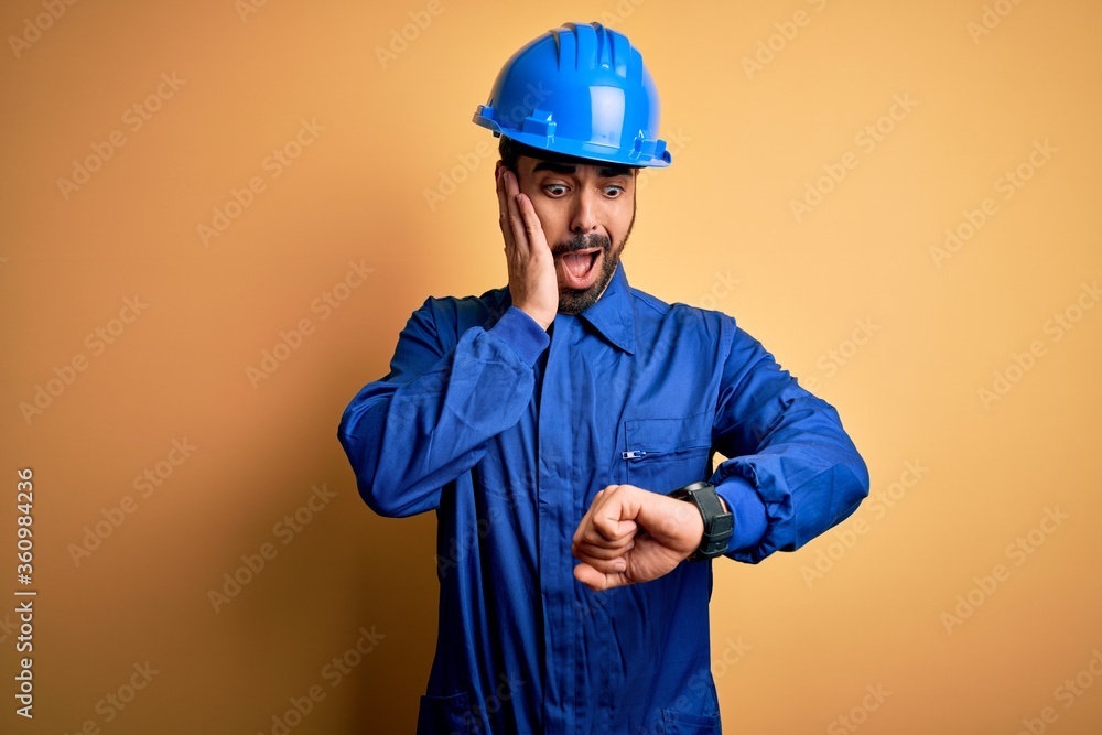 Mechanic man with beard wearing blue uniform and safety helmet over yellow background Looking at the watch time worried, afraid of getting late