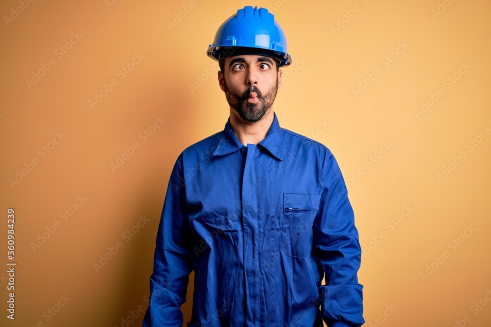 Mechanic man with beard wearing blue uniform and safety helmet over yellow background making fish face with lips, crazy and comical gesture. Funny expression.