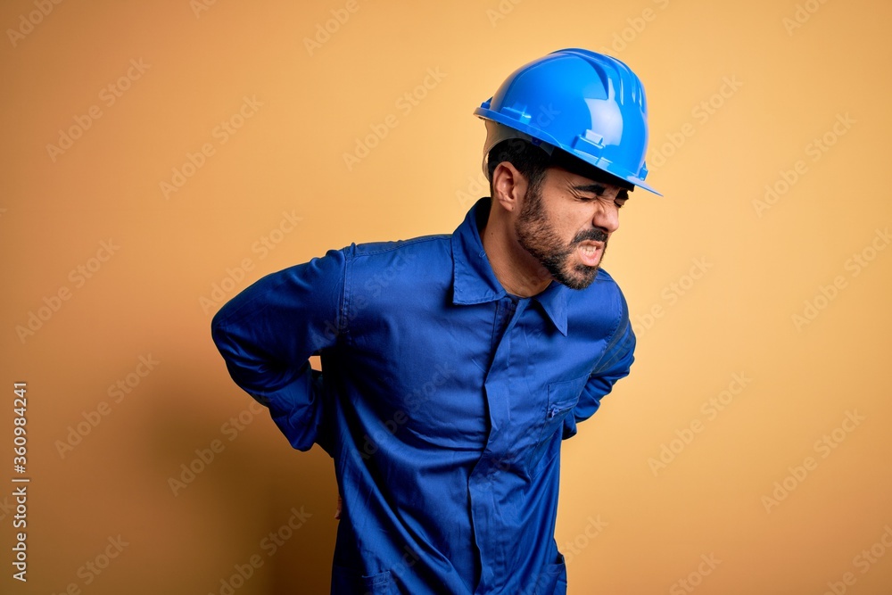 Mechanic man with beard wearing blue uniform and safety helmet over yellow background Suffering of backache, touching back with hand, muscular pain