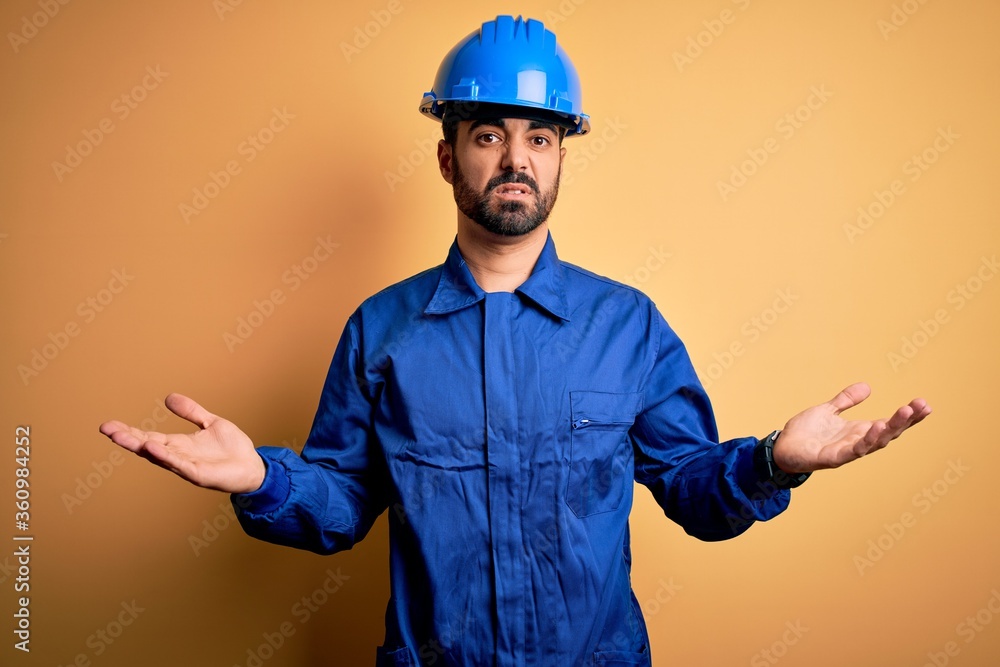 Mechanic man with beard wearing blue uniform and safety helmet over yellow background clueless and confused with open arms, no idea concept.