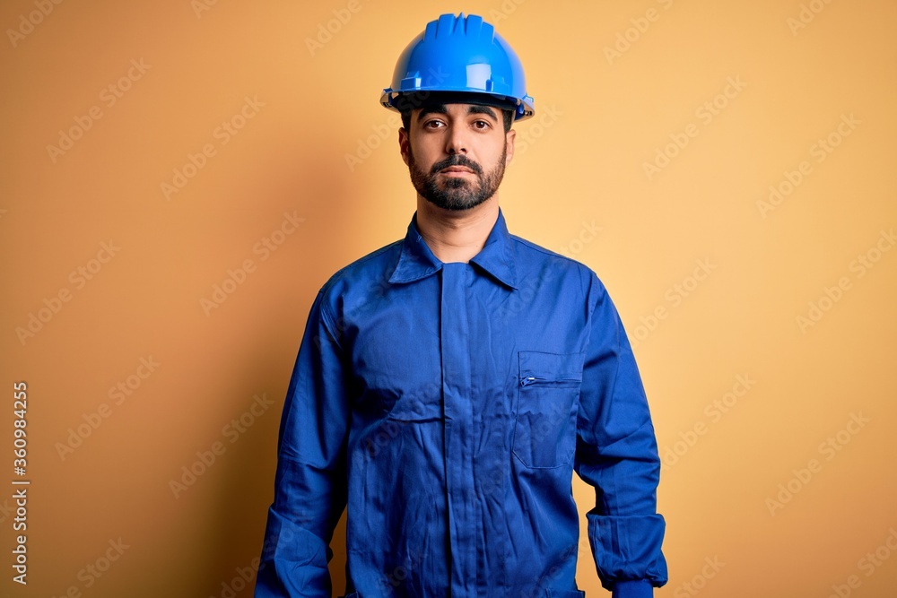 Mechanic man with beard wearing blue uniform and safety helmet over yellow background Relaxed with serious expression on face. Simple and natural looking at the camera.