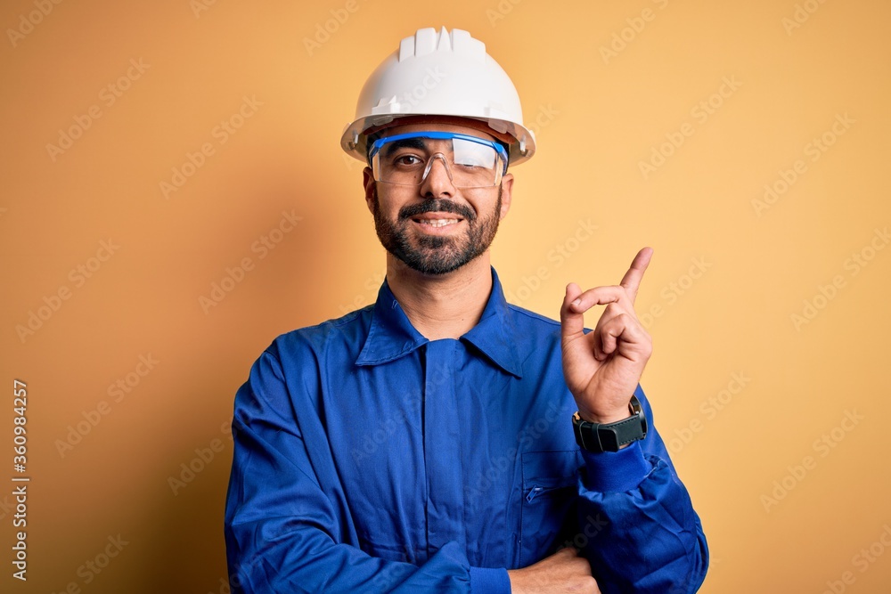 Mechanic man with beard wearing blue uniform and safety glasses over yellow background with a big smile on face, pointing with hand and finger to the side looking at the camera. Mechanic man with beard wearing blue uniform and safety glasses over yellow background with a big smile on face, pointing with hand and finger to the side looking at the camera.
