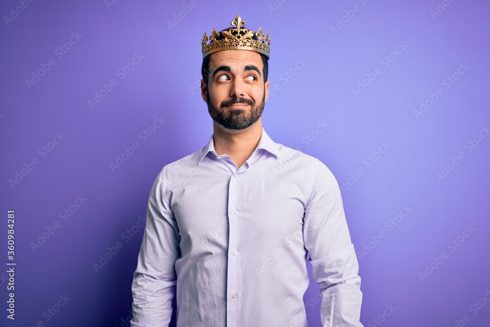 Young handsome man with beard wearing golden crown of king over purple background smiling looking to the side and staring away thinking. Young handsome man with beard wearing golden crown of king over purple background smiling looking to the side and staring away thinking.