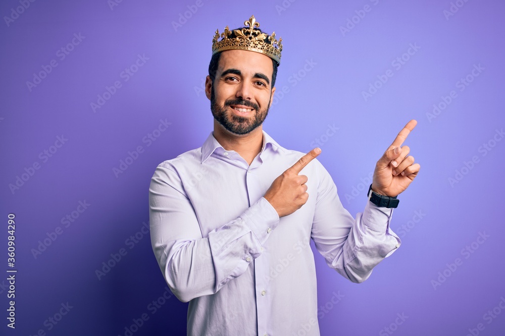 Young handsome man with beard wearing golden crown of king over purple background smiling and looking at the camera pointing with two hands and fingers to the side. Young handsome man with beard wearing golden crown of king over purple background smiling and looking at the camera pointing with two hands and fingers to the side.