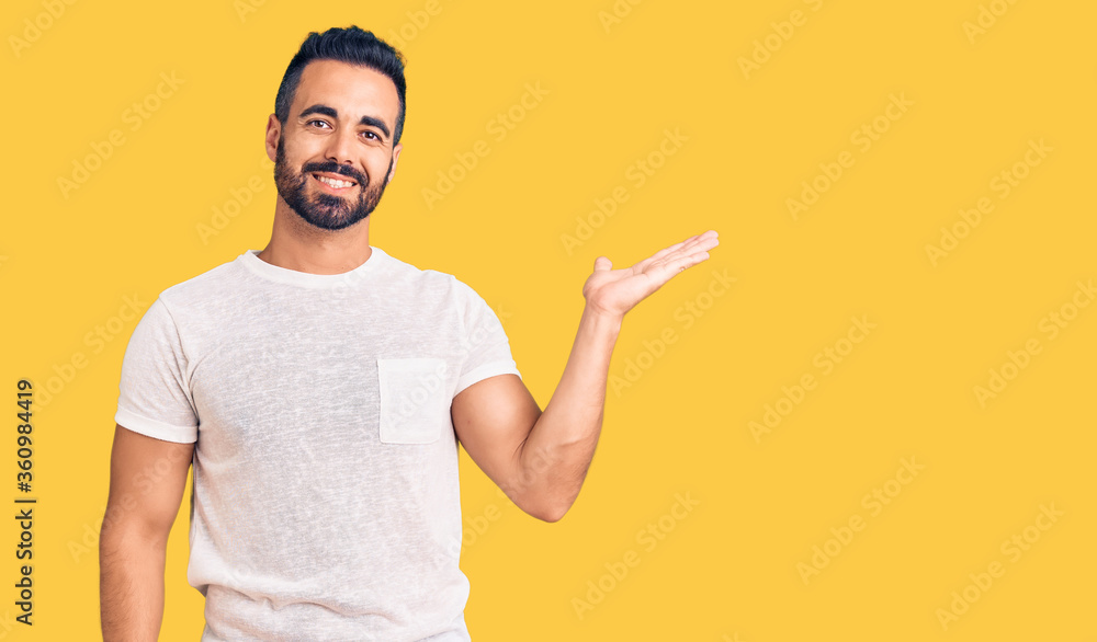 Young hispanic man wearing casual clothes smiling cheerful presenting and pointing with palm of hand looking at the camera. Young hispanic man wearing casual clothes smiling cheerful presenting and pointing with palm of hand looking at the camera.