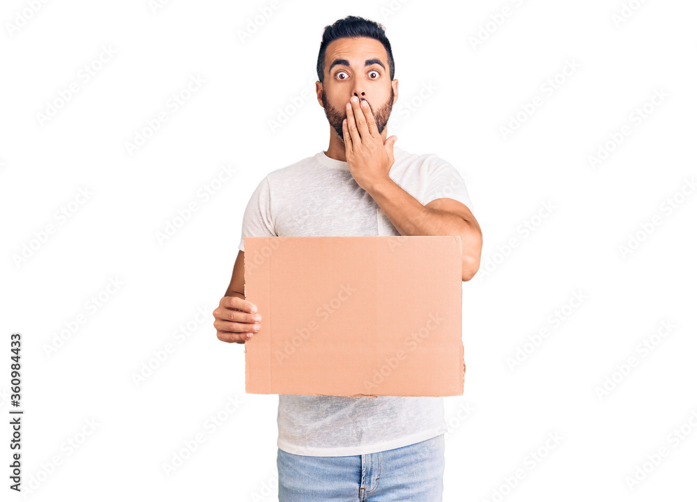 Young hispanic man holding banner cardboard covering mouth with hand, shocked and afraid for mistake. surprised expression Young hispanic man holding banner cardboard covering mouth with hand, shocked and afraid for mistake. surprised expression