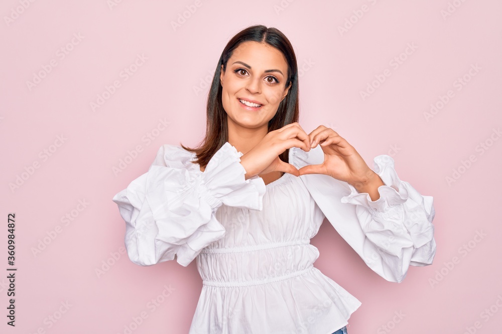 Young beautiful brunette woman wearing casual white dress standing over pink background smiling in love showing heart symbol and shape with hands. Romantic concept. Young beautiful brunette woman wearing casual white dress standing over pink background smiling in love showing heart symbol and shape with hands. Romantic concept.