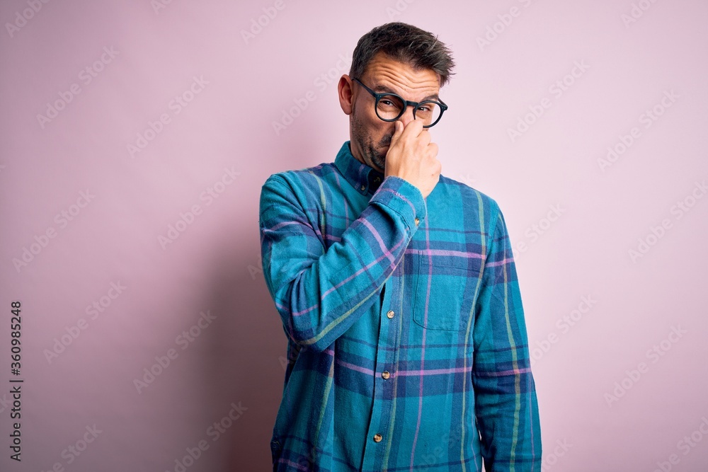 Young handsome man wearing casual shirt and glasses standing over isolated pink background smelling something stinky and disgusting, intolerable smell, holding breath with fingers on nose. Bad smell Young handsome man wearing casual shirt and glasses standing over isolated pink background smelling something stinky and disgusting, intolerable smell, holding breath with fingers on nose. Bad smell