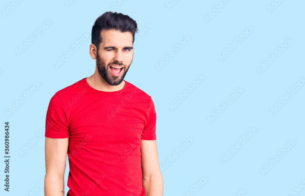 Young handsome man with beard wearing casual t-shirt winking looking at the camera with sexy expression, cheerful and happy face. Young handsome man with beard wearing casual t-shirt winking looking at the camera with sexy expression, cheerful and happy face.