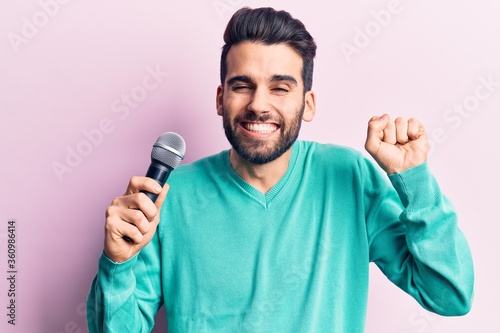 Young handsome man with beard singing song using microphone screaming proud, celebrating victory and success very excited with raised arm