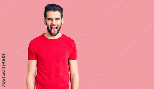 Young handsome man with beard wearing casual t-shirt sticking tongue out happy with funny expression. emotion concept.
