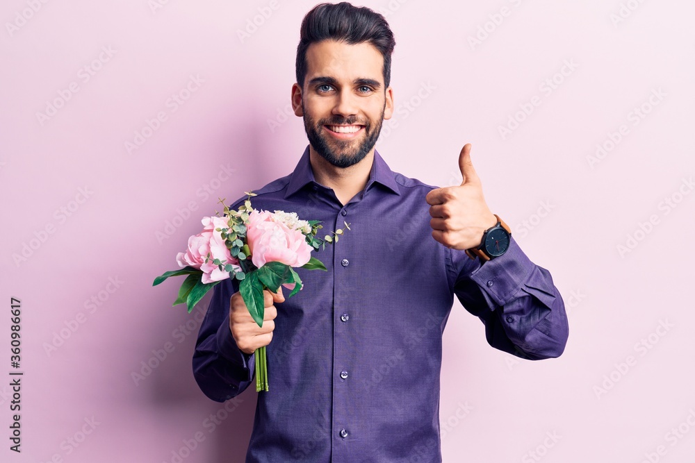 Young handsome man with beard holding bouquet of flowers smiling happy and positive, thumb up doing excellent and approval sign