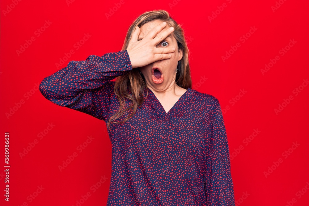 Young beautiful blonde woman standing over red isolated background peeking in shock covering face and eyes with hand, looking through fingers afraid Young beautiful blonde woman standing over red isolated background peeking in shock covering face and eyes with hand, looking through fingers afraid