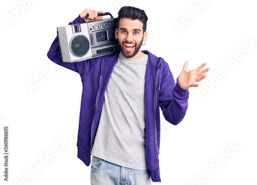 Young handsome man with beard listening to music using vintage boombox celebrating victory with happy smile and winner expression with raised hands