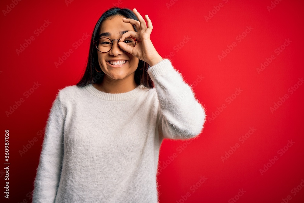 Young beautiful asian woman wearing casual sweater and glasses over red background doing ok gesture with hand smiling, eye looking through fingers with happy face. Young beautiful asian woman wearing casual sweater and glasses over red background doing ok gesture with hand smiling, eye looking through fingers with happy face.