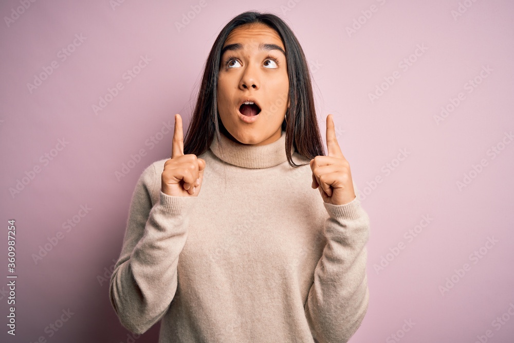Young beautiful asian woman wearing casual turtleneck sweater over pink background amazed and surprised looking up and pointing with fingers and raised arms. Young beautiful asian woman wearing casual turtleneck sweater over pink background amazed and surprised looking up and pointing with fingers and raised arms.