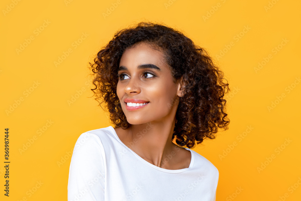 Close-up portrait of smiling young beautiful African American woman thinking while happily looking at the side in isolated studio yellow background Close-up portrait of smiling young beautiful African American woman thinking while happily looking at the side in isolated studio yellow background
