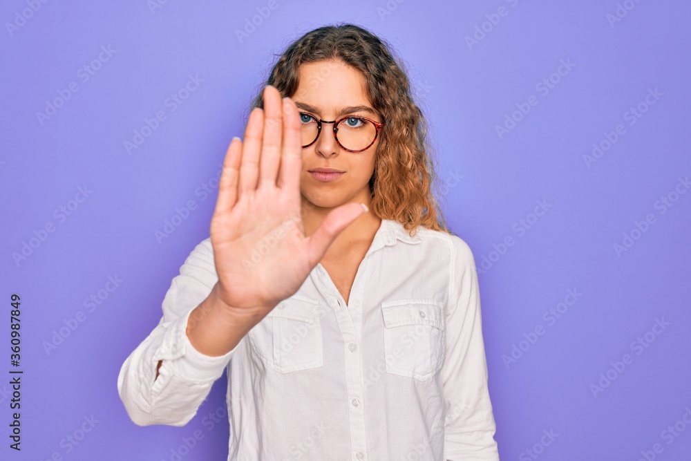 Young beautiful woman with blue eyes wearing casual shirt and glasses over purple background doing stop sing with palm of the hand. Warning expression with negative and serious gesture on the face. Young beautiful woman with blue eyes wearing casual shirt and glasses over purple background doing stop sing with palm of the hand. Warning expression with negative and serious gesture on the face.