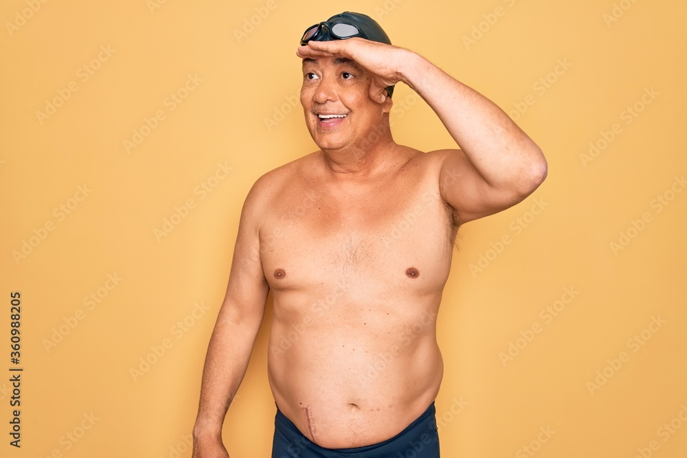 Middle age senior grey-haired swimmer man wearing swimsuit, cap and goggles very happy and smiling looking far away with hand over head. Searching concept.