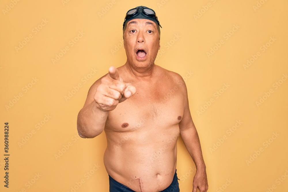 Middle age senior grey-haired swimmer man wearing swimsuit, cap and goggles pointing displeased and frustrated to the camera, angry and furious with you