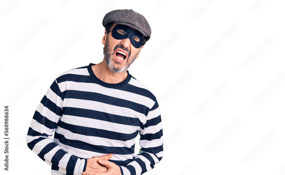Middle age handsome man wearing burglar mask with hand on stomach because nausea, painful disease feeling unwell. ache concept. Middle age handsome man wearing burglar mask with hand on stomach because nausea, painful disease feeling unwell. ache concept.