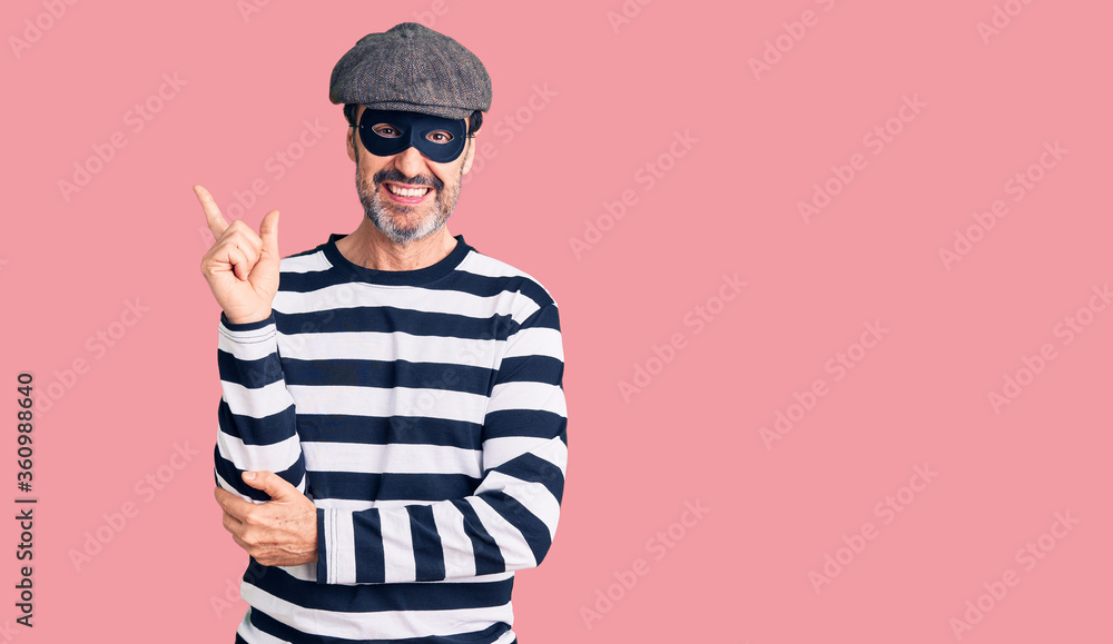 Middle age handsome man wearing burglar mask with a big smile on face, pointing with hand and finger to the side looking at the camera. Middle age handsome man wearing burglar mask with a big smile on face, pointing with hand and finger to the side looking at the camera.