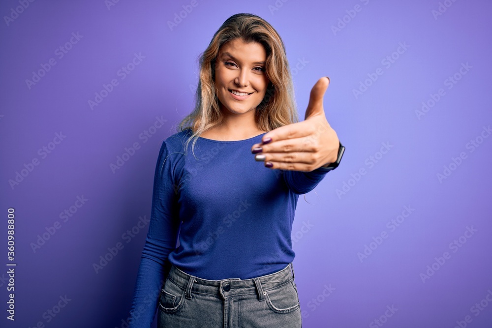 Young beautiful blonde woman wearing casual t-shirt over isolated purple background smiling friendly offering handshake as greeting and welcoming. Successful business. Young beautiful blonde woman wearing casual t-shirt over isolated purple background smiling friendly offering handshake as greeting and welcoming. Successful business.