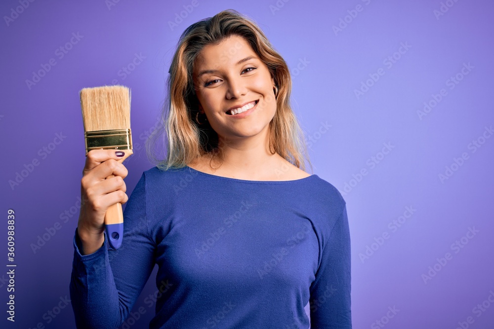 Young beautiful blonde painter woman painting using paint bush over purple background with a happy face standing and smiling with a confident smile showing teeth Young beautiful blonde painter woman painting using paint bush over purple background with a happy face standing and smiling with a confident smile showing teeth