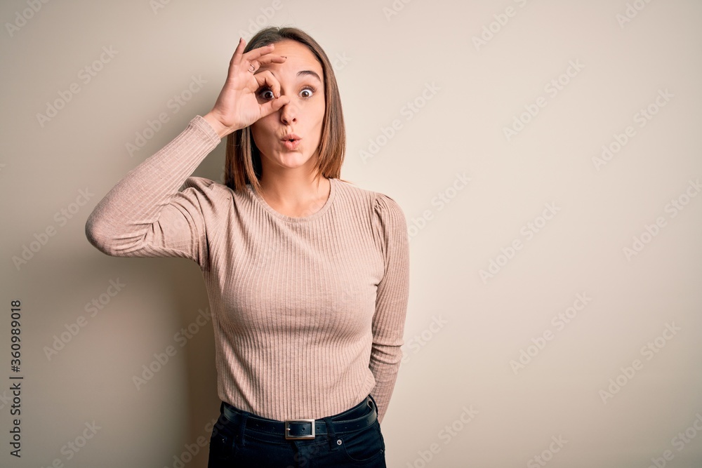 Young beautiful woman wearing casual sweater standing over isolated white background doing ok gesture shocked with surprised face, eye looking through fingers. Unbelieving expression. Young beautiful woman wearing casual sweater standing over isolated white background doing ok gesture shocked with surprised face, eye looking through fingers. Unbelieving expression.