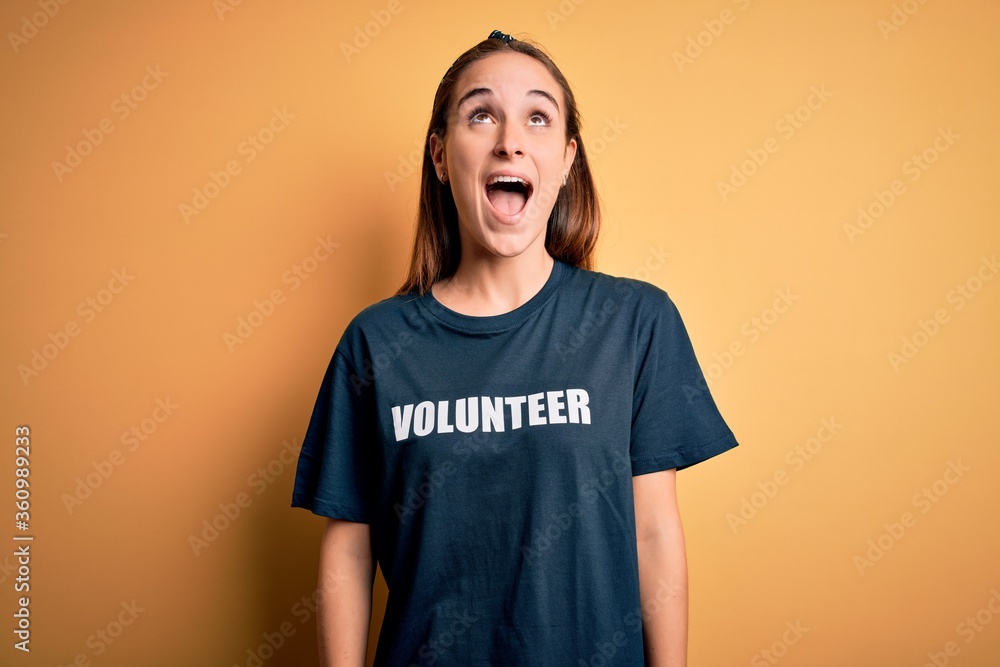 Young beautiful woman wearing volunteer t-shirt doing volunteering over yellow background angry and mad screaming frustrated and furious, shouting with anger. Rage and aggressive concept. Young beautiful woman wearing volunteer t-shirt doing volunteering over yellow background angry and mad screaming frustrated and furious, shouting with anger. Rage and aggressive concept.