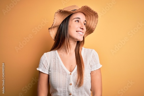 Young beautiful woman wearing casual t-shirt and summer hat over isolated yellow background looking away to side with smile on face, natural expression. Laughing confident.