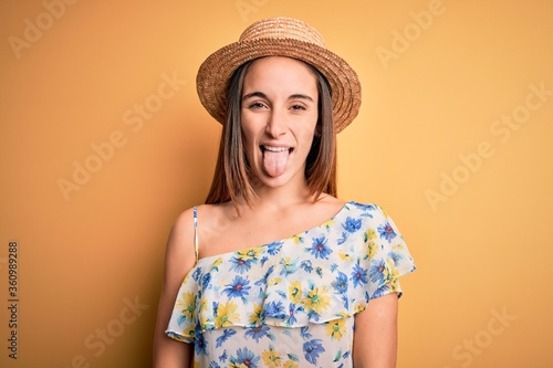 Young beautiful woman wearing casual t-shirt and summer hat over isolated yellow background sticking tongue out happy with funny expression. Emotion concept.