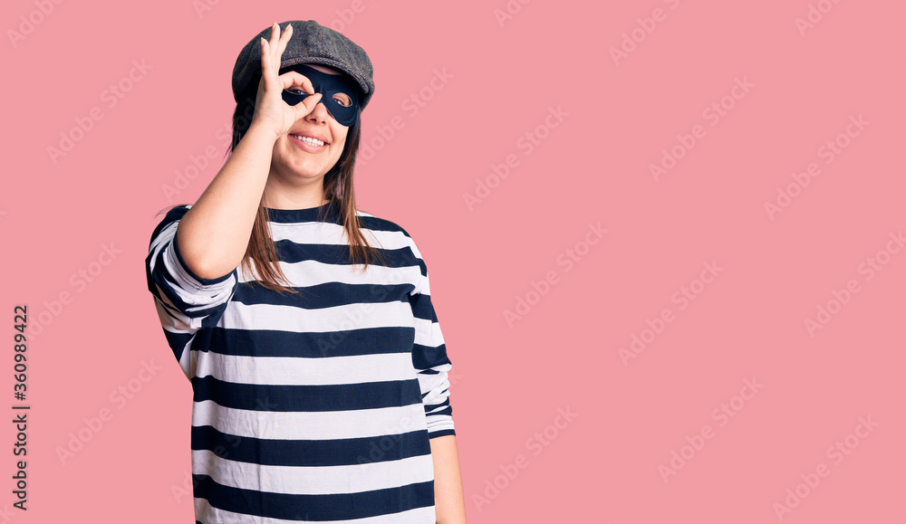 Young beautiful brunette woman wearing burglar mask doing ok gesture with hand smiling, eye looking through fingers with happy face. Young beautiful brunette woman wearing burglar mask doing ok gesture with hand smiling, eye looking through fingers with happy face.