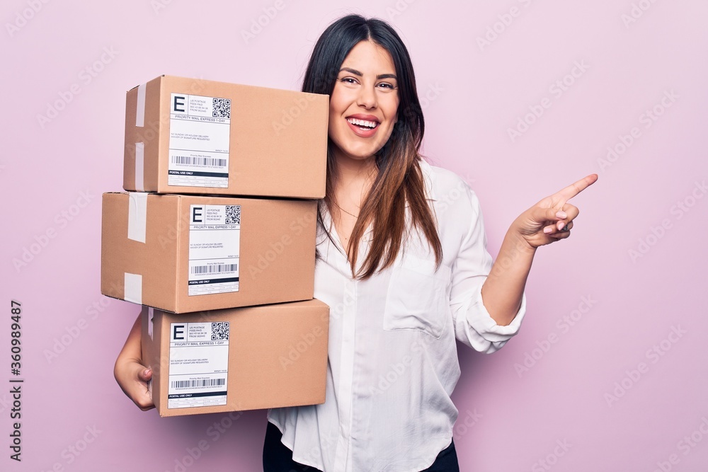 Young beautiful brunette woman holding deliver packages standing over pink background smiling happy pointing with hand and finger to the side Young beautiful brunette woman holding deliver packages standing over pink background smiling happy pointing with hand and finger to the side