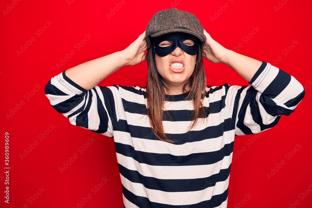 Young beautiful brunette burglar woman wearing cap and mask over isolated red background with hand on head, headache because stress. Suffering migraine. Young beautiful brunette burglar woman wearing cap and mask over isolated red background with hand on head, headache because stress. Suffering migraine.