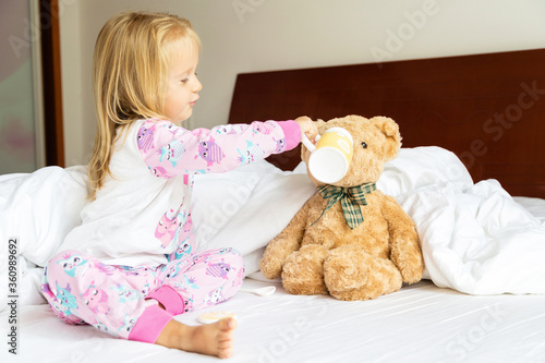 Cute little girl with blonde hair sitting on the bed with stuffed teddy bear. Happy childhood. Stay at home during coronavirus covid-19 pandemic quarantine concept.