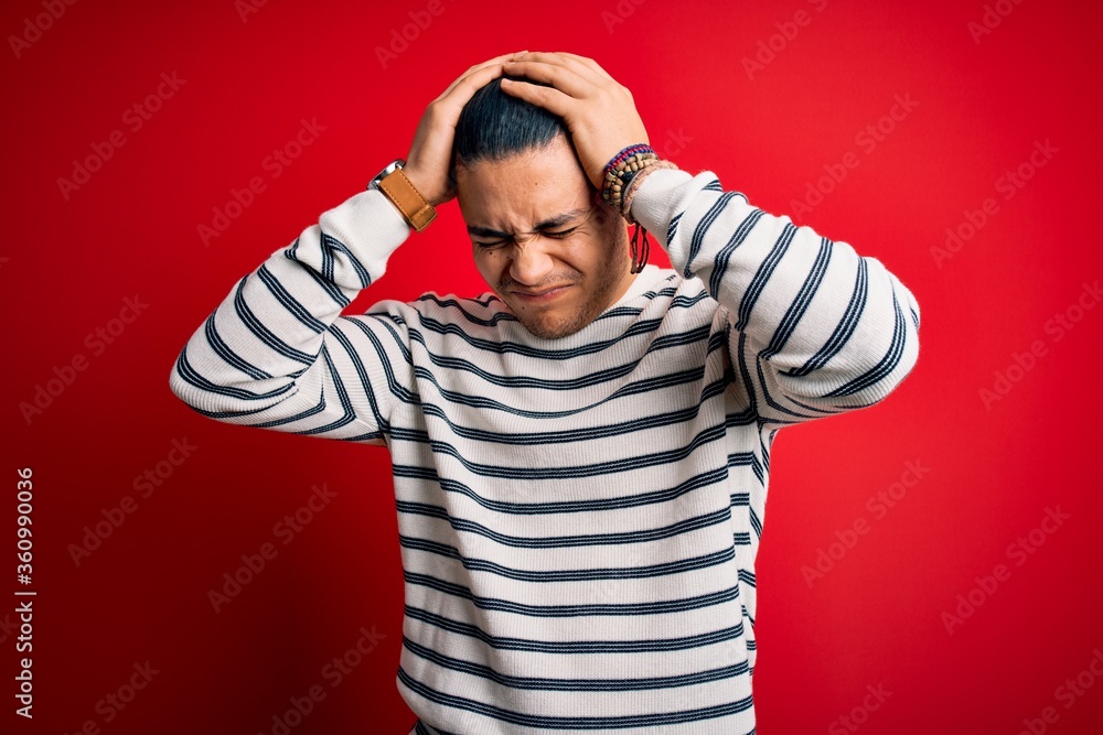 Young brazilian man wearing casual striped t-shirt standing over isolated red background suffering from headache desperate and stressed because pain and migraine. Hands on head. Young brazilian man wearing casual striped t-shirt standing over isolated red background suffering from headache desperate and stressed because pain and migraine. Hands on head.