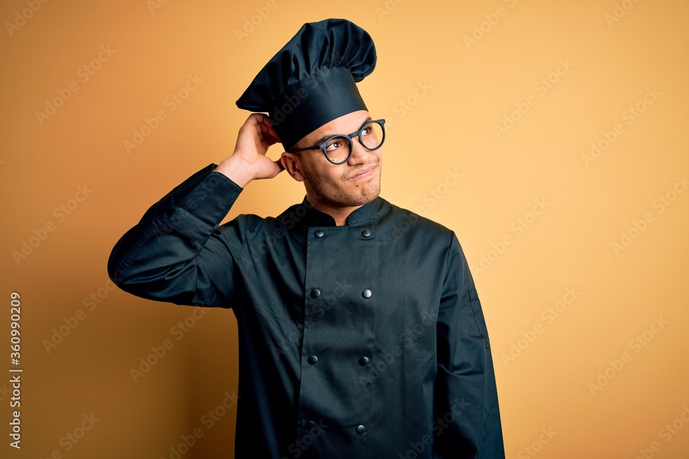 Young brazilian chef man wearing cooker uniform and hat over isolated yellow background confuse and wondering about question. Uncertain with doubt, thinking with hand on head. Pensive concept. Young brazilian chef man wearing cooker uniform and hat over isolated yellow background confuse and wondering about question. Uncertain with doubt, thinking with hand on head. Pensive concept.