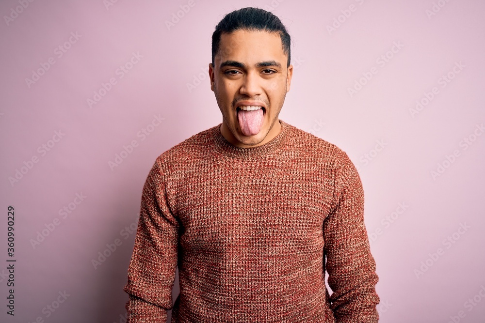 Young brazilian man wearing casual sweater standing over isolated pink background sticking tongue out happy with funny expression. Emotion concept. - obrazy, fototapety, plakaty 
