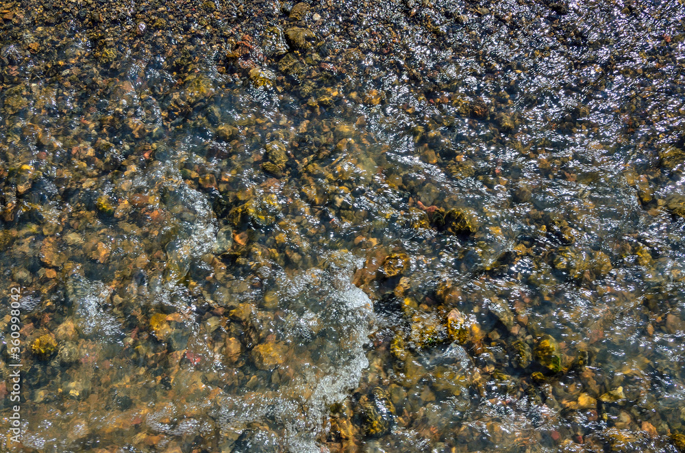 Cool freshness of mountain river fast stream in sunny summer day. Colorful pebbles under transparent clear water, sun glare on the surface of  water 